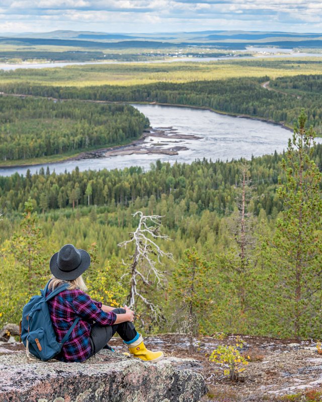 Värikkäästi pukeutunut retkeilijä istuu ja ihailee Väylänvarren jokimaisemaa.